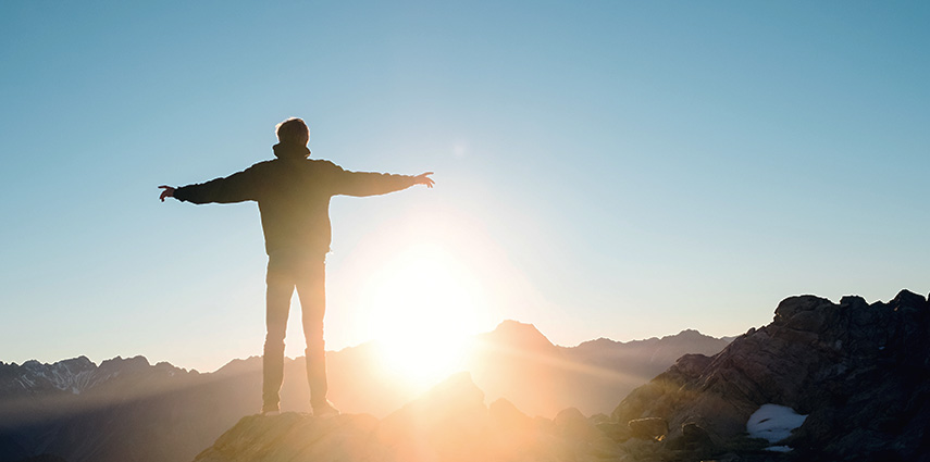 Homem no topo da montanha, braços abertos ao horizonte, ao nascer do Sol. Tranquilidade, resiliência e superação.